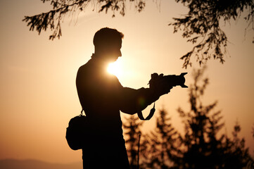 Silhouette of a photographer in the mountains taking pictures at dawn. Tourist holding camera on the mountain hill on sunrise background. Man takes photo of mountain in the rays of the setting sun.