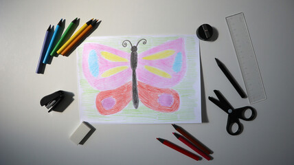 Photo of child's drawing with colored pencils. In the picture is butterfly. Office equipment around. Colored pencils, scissors, sharpener, ruler, etc. Pink, red, blue, green colors. White background.