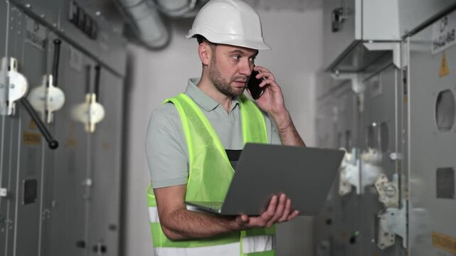 Professional electrical engineer in hard hat and reflective vest diligently works in an electrical control room. He carefully reviews information on a laptop while having a serious phone conversation.