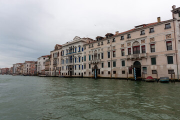 A panoramic view of the Grand Canal in Venice, Italy, showcasing the ornate facades of historic palazzi reflecting in the water. Several boats can be seen navigating the canal. Overcast sky