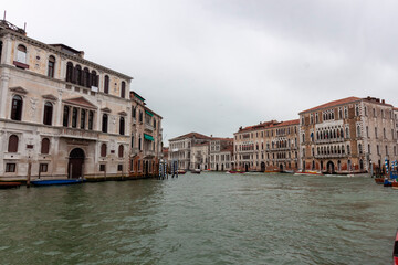 A panoramic view of the Grand Canal in Venice, Italy, showcasing the ornate facades of historic palazzi reflecting in the water. Several boats can be seen navigating the canal. Overcast sky