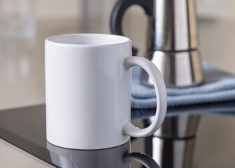 White coffee mug on induction stove against moka pot on blue dish cloth closeup, mockup