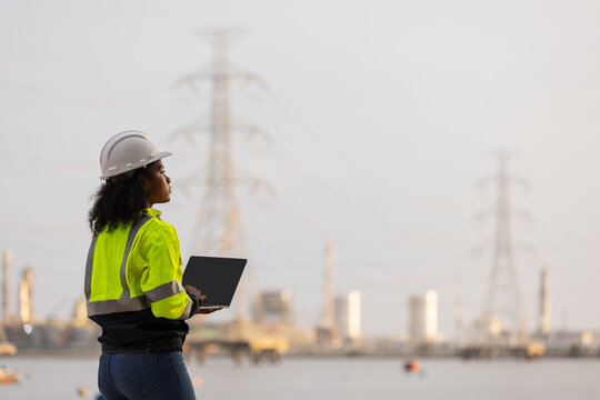 A female engineer in a high-visibility jacket and helmet uses a laptop and smartphone near a power plant and transmission towers, illustrating modern technology, communication, and industrial work