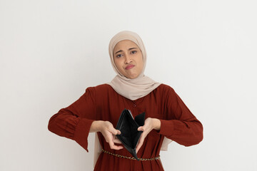 Obraz premium Sad young woman in a hijab looking up while holding an empty wallet, expressing financial difficulties or disappointment.