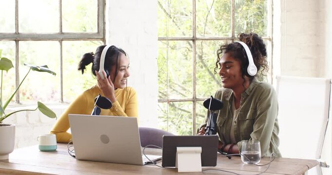 Two multiracial female colleagues recording podcast at home, with headphones and using microphones