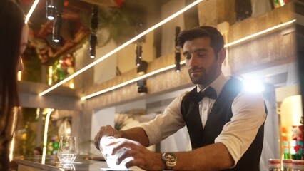 Focusing on smart bartender serving gin fizz or tonic at luxurious bar while engaging lively conversation with VIP customer at back view in lighting atmosphere at lighting warm nightclub. Vinosity.