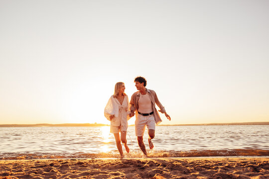Smiling beautiful woman and her handsome boyfriend. Excited couple in casual clothes. Happy cheerful family. Female and man run at sunrise over sea beach outdoors. Run at seaside in summer day