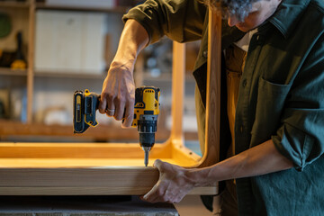 Carpenter attaches wooden table base to tabletop using power drill in joinery workshop. Handyman...