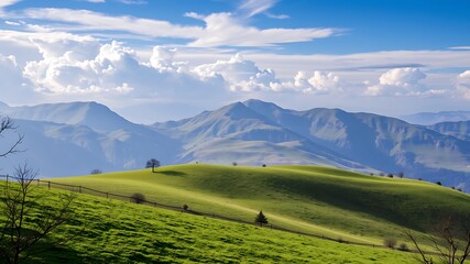 Obraz premium Serene spring landscape: rolling green hills and majestic mountains under a blue sky with fluffy clouds, perfect for nature and travel photography.