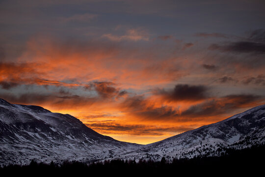 Vall&eacute;e enneig&eacute;e et bois&eacute;e entre deux montagnes sous un ciel orange cr&eacute;pusculaire et nuageux du soleil couchant en hiver.