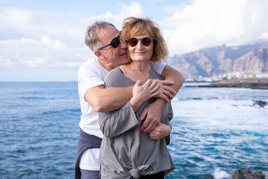 Happy mature and romantic senior couple exchange cuddles and hug each other on the seashore in a sunny day expressing love and tenderness