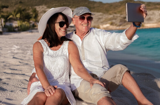 Smiling senior couple sitting at the beach make a selfie with digital tablet enjoying the sunset expressing love, happiness, and relax. Vacation, travel, retirement concept