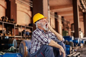 Tired senior industry worker with helmet on head yawning while sitting at facility.