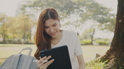 Obraz premium Asian Woman Working Remotely in a Park, Using Tablet for Hybrid Work Lifestyle, Tranquil Freelance, Joyful Digital Nomad in Nature, Relaxing and Working in Nature, Remote work using Tablet in greenery