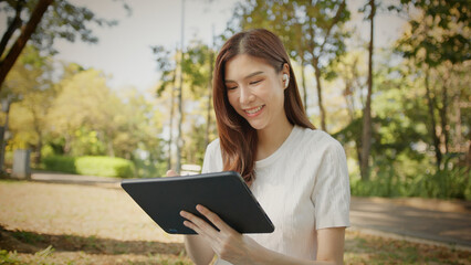 Fototapeta premium Asian Woman Working outdoors in the park. Professional on a Video Call, exploring Hybrid Work Opportunities in a Peaceful, Virtual Meeting and Online Learning. Hybrid work lifestyle concept by tablet