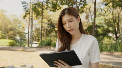 Asian Woman Working Remotely in a Park, Using Tablet for Hybrid Work Lifestyle, Tranquil Freelance, Joyful Digital Nomad in Nature, Relaxing and Working in Nature, Remote work using Tablet in greenery