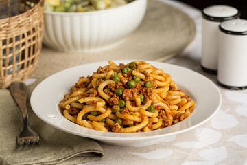 Plate with italian pasta gramigna with meat and tomato ragu sauce and green peas. Table served for lunch, salad and bottle of water on background.
