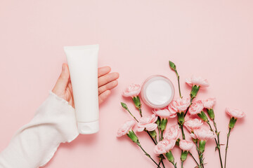 Woman hand hold mockup bottle facial cream with pink flowers on gentle pink background, top view. Skincare set, cute girlish concept