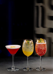A trio of colorful cocktails in elegant glassware garnished with fruits and herbs on a dark background.