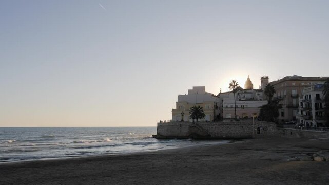 Beautiful sunset in seaside medieval Sitges city. Church of Sant Bartolomeu i Santa Tecla. Golden hour colours. Sun reflecting on the buildings. 