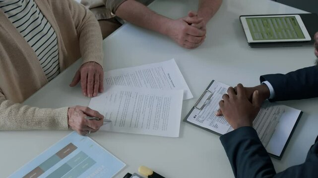 Close-up view of Caucasian senior client signing mortgage agreement while Black financial advisor pointing at signature block in document during office meeting