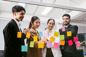 A diverse team collaborates around a glass wall covered in colorful sticky notes, brainstorming...