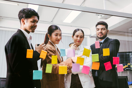 A diverse team collaborates around a glass wall covered in colorful sticky notes, brainstorming ideas in a modern office setting. Using agile methodology for business in a tech start-up office.