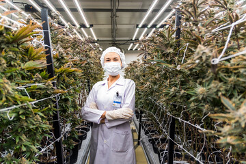 A woman in protective gear stands confidently among cannabis plants in a well-lit indoor cultivation facility.