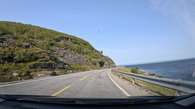 Driving through the tundra on E69 in northern Norway. Rocky hills with sparse trees and grass rise on the left, while the sea stretches along the right, showcasing raw and rugged nature.