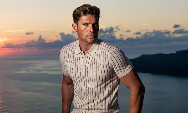 A man in a short-sleeved polo shirt and beige chinos leans against a railing, looking out over the sunset sea. 