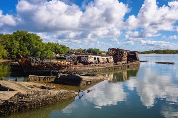 A rusty shipwreck in calm waters with lush greenery and a blue sky.
