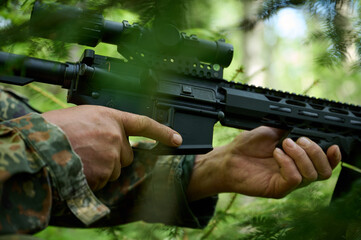 Hunter with rifle wearing camouflage suit sitting in ambush. Camouflaged sniper in the forest in...
