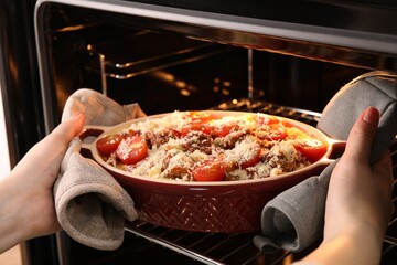 Woman putting pasta casserole with cheese, tomatoes and minced meat into oven, closeup