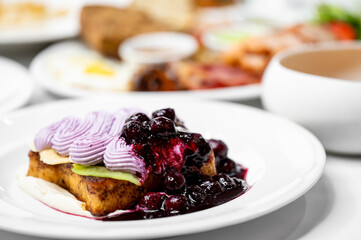 A close-up of a delicious dessert featuring a slice of cake topped with creamy purple frosting and a berry compote, served on a white plate, with a blurred background of various dishes.