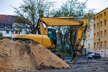 A yellow excavator stands behind a large pile of clay against the background of houses and cars.