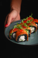 Hands of a girl with red nails holding a plate of sushi rolls on a dark background