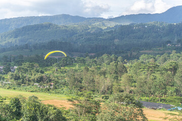 A yellow paraglider soars above lush, green hills with cloudy skies in the background.