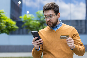 A young serious man is standing on the street, holding a credit card, looking at the mobile phone...