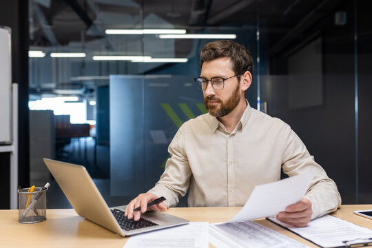 A concentrated young man is sitting in the office at a desk, holding documents in his hands and working on a laptop