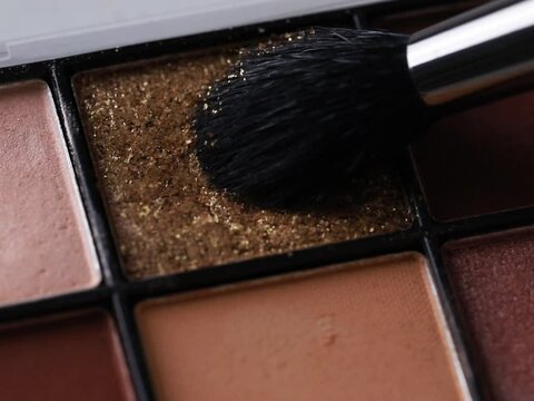 A close-up image of an eyeshadow palette showcasing a shimmery, textured golden brown shade in focus, with a makeup brush gently hovering over it