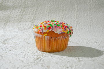 A colorful cupcake topped with vanilla frosting and a generous amount of rainbow sprinkles, set against a textured white background for a clean and appealing look