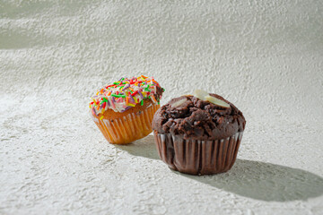 Two cupcakes on a textured white background. vanilla cupcake with white frosting covered in rainbow sprinkles and chocolate cupcakes showcasing a variety of sweet treats