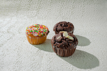 three of cupcakes on textured white: one with rainbow sprinkles, one chocolate with chocochips, and one chocolate with almond slices