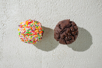 Two cupcakes on a textured white background. vanilla cupcake with white frosting covered in rainbow sprinkles and chocolate cupcakes showcasing a variety of sweet treats