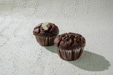 Two of cupcakes on textured white: one chocolate with chocochips and one chocolate with almond slices