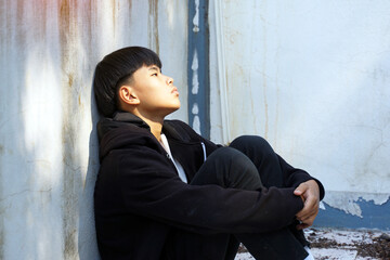 teenage boy sits, hugging his knees with his back against the wall of an abandoned building, alone. Concept of lonely, abandoned, quarantined, depressed.