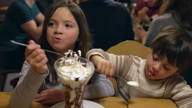 Kdis eating whipped cream sundae at restaurant, brother and sister sharing sugar treat indulgence at diner in the evening weekend outing