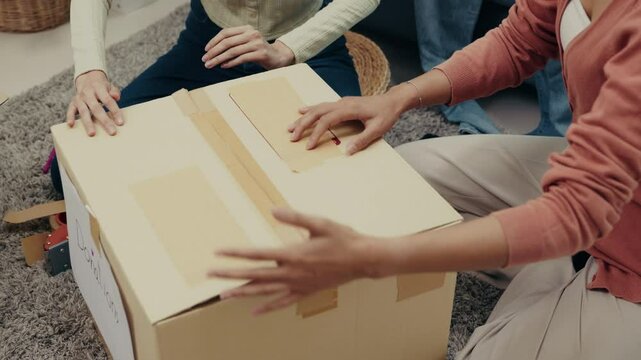 Two Asian women work together to seal a donation box with tape, preparing clothes and household items for charity, promoting generosity and sustainability.