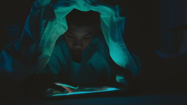 A boy is sitting in a dark room with a tablet in his hand. He is looking at the screen with a serious expression