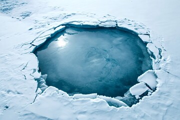 Melting ice creating a deep blue water pool surrounded by thick snow and ice on a frozen lake during winter, creating a beautiful contrast between the cold white snow and the deep blue water